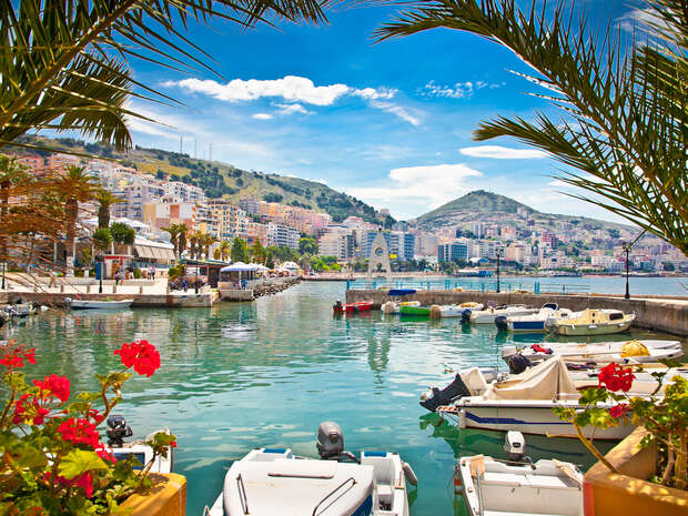 Boats float on a calm, turquoise harbor, framed by palm fronds and vibrant flowers, with a hillside town and cloud-dotted sky in the background.